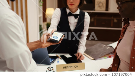 Receptionist using POS payment system to validate card purchase of extra room amenities and hotel services, close up. African american couple using smartphone banking app during booking in process Receptionist using POS payment system to validate card purchase of extra room amenities and hotel services, close up. African american couple using smartphone banking app during booking in process 107197004