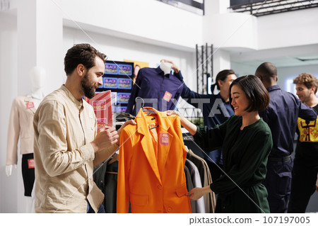 Happy diverse couple shopping on Black Friday in clothing store, looking at orange formal jacket with red sale tag and discussing price. Husband helping wife to choose clothes during seasonal sales Happy diverse couple shopping on Black Friday in clothing store, looking at orange formal jacket with red sale tag and discussing price. Husband helping wife to choose clothes during seasonal sales 107197005