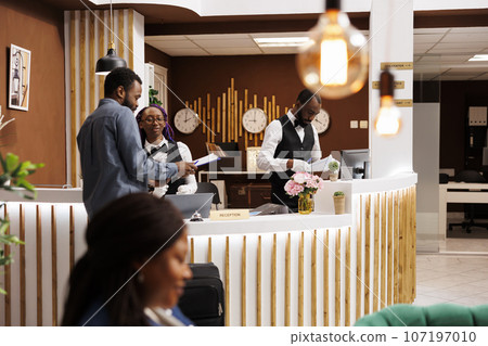 African American man tourist standing at reception with luggage filling check-in form upon arrival at hotel. Black guy traveler signing documents at front desk while checking in African American man tourist standing at reception with luggage filling check-in form upon arrival at hotel. Black guy traveler signing documents at front desk while checking in 107197010
