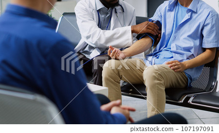 Cardiologist using tonometer instrument to measure patient pulse pressure and hypertension in hospital waiting room. African american doctor doing cardiology exam with asian man during checkup visit Cardiologist using tonometer instrument to measure patient pulse pressure and hypertension in hospital waiting room. African american doctor doing cardiology exam with asian man during checkup visit 107197091