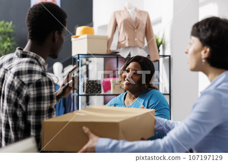 Stylish worker giving carton box to african american customer, standing at counter desk in clothing store. Shopaholic man buying fashionable merchandise in modern boutique. Fashion concept 107197129