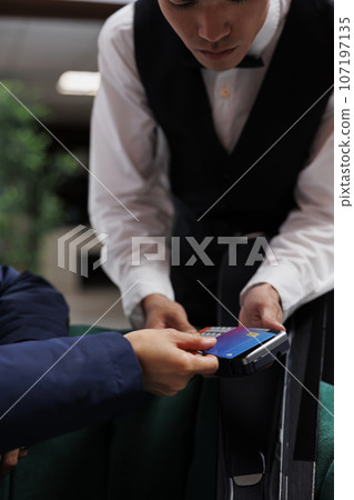 Close up of male concierge grasping pos machine and facing customer paying with debit card in hotel lobby. In lounge area of the ski resort a detailed view of credit card used for cashless payments. Close up of male concierge grasping pos machine and facing customer paying with debit card in hotel lobby. In lounge area of the ski resort a detailed view of credit card used for cashless payments. 107197135