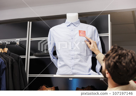 Male customer checking price on shirt while standing in clothing store, looking at red sale tag sticker. Man shopper counting discount while making purchases in clothes store on Black Friday Male customer checking price on shirt while standing in clothing store, looking at red sale tag sticker. Man shopper counting discount while making purchases in clothes store on Black Friday 107197146