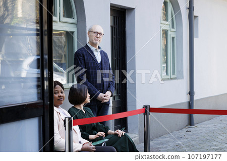Diverse people customers sitting outdoors at clothing store entrance in early morning, waiting for Black Friday deals on city street. Shoppers lined up outside of shopping mall 107197177