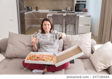 Girl eating pizza sitting on couch and watching tv in modern appartment Girl eating pizza sitting on couch and watching tv in modern appartment 107198848