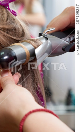 Hairdresser makes hairstyle girl with long hair in a beauty salon. Create curls with curling irons Hairdresser makes hairstyle girl with long hair in a beauty salon. Create curls with curling irons 107199287