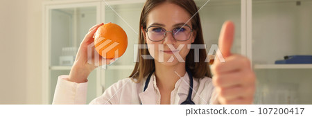 Young nutritionist holds orange fruit and shows thumb up. 107200417