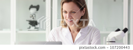 Satisfied female researcher working on laptop at laboratory table. 107200443