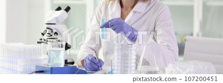 Smiling female scientist holding test tube with blue liquid and making notes. 107200451