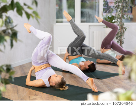 Yoga class of sporty women in activewear performing Halasana or Plow Pose on mats during workout in cozy yoga studio Yoga class of sporty women in activewear performing Halasana or Plow Pose on mats during workout in cozy yoga studio 107204846
