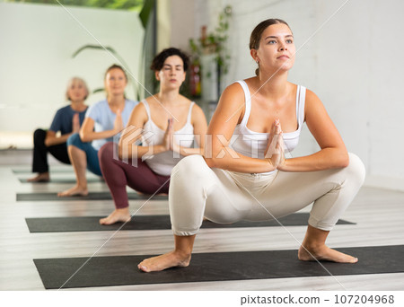 Calm European young woman standing Squat Pose Malasana during yoga practice in fitness class Calm European young woman standing Squat Pose Malasana during yoga practice in fitness class 107204968