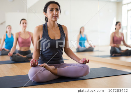 Concentrated Latina sitting in lotus pose with group of women Concentrated Latina sitting in lotus pose with group of women 107205422