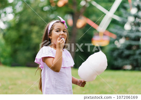 Child girl in an amusement park in the summer eats cotton candy and smiles happily. The concept of summer holidays and school holidays 107206169