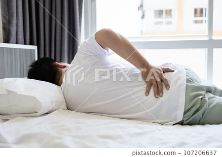 Stressed asian man having a backache,sore hips,waist hurts,unhappy adult people suffering from low lumbar pain or acute back strain,lying on the mattress at home,health care,medical,lifestyle concept 107206637