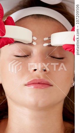 Beautiful Woman in Spa Clinic Receiving Stimulating Electric Facial Treatment From Therapist. Closeup Of Young Female Face During Microcurrent Therapy 107209506