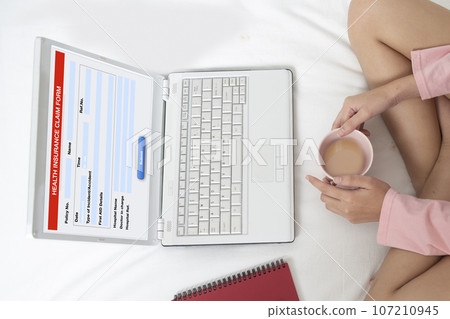 Young woman sits on the bed and fills out electronic health insurance claim form. 107210945