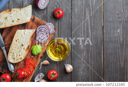 Fresh baguette sliced into pieces, tomatoes, vegetables and olive oil, ingredients for making a vegan sandwich, close-up. Dark wooden background, copy space for text Fresh baguette sliced into pieces, tomatoes, vegetables and olive oil, ingredients for making a vegan sandwich, close-up. Dark wooden background, copy space for text 107212395