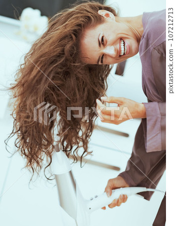 Smiling young woman blow drying hair in bathroom Smiling young woman blow drying hair in bathroom 107212713