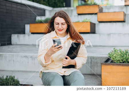 Elegant young 30s woman formally dressed, sitting on the bench and holding a folder with documents outdoors. Businesswoman with smart phone standing at the city. Elegant young 30s woman formally dressed, sitting on the bench and holding a folder with documents outdoors. Businesswoman with smart phone standing at the city. 107213189