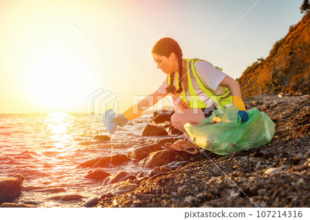 Side view of young squatting volunteer wearing rubber gloves collects garbage on wild coast. Concept of environment disaster and coast cleanup 107214316