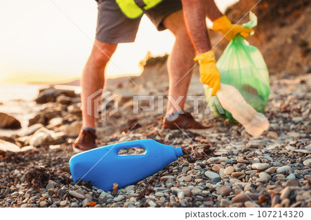 Close up view of walking volunteer holding full plastic bag at pebble wild beach. Close up of plastic bottle in foreground. Concept of protection environment and Earth Day Close up view of walking volunteer holding full plastic bag at pebble wild beach. Close up of plastic bottle in foreground. Concept of protection environment and Earth Day 107214320