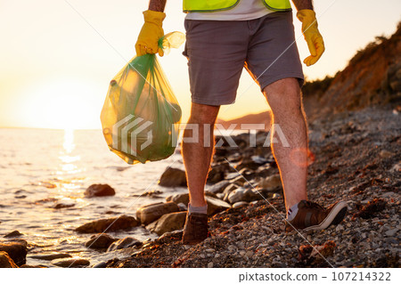 Close up of male legs of volunteer walking on wild coast holding plastic bag full of garbage. Concept of ecological disaster and volunteering clean up Close up of male legs of volunteer walking on wild coast holding plastic bag full of garbage. Concept of ecological disaster and volunteering clean up 107214322