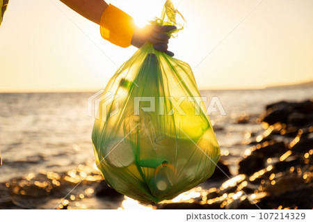 Close up of hand wearing glove and holds bog plastic full trash bag. Ocean in background. Concept of ecological disaster and ocean cleanup 107214329