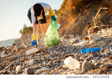 Coast pollution. Low angle view of young Caucasian woman in rubber gloves collects garbage on pebble wild beach. Concept of save Earth and environment disaster 107214339