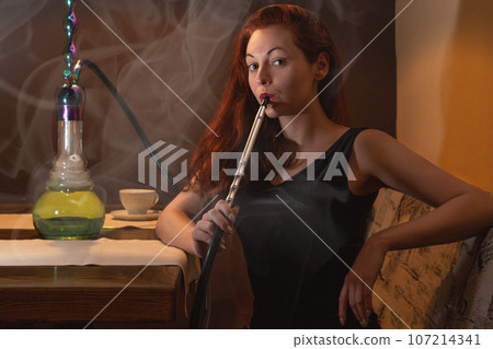 Young caucasian woman smokes a hookah or shisha in the night club or bar smoke. Smoke and steam in the air. Close up Young caucasian woman smokes a hookah or shisha in the night club or bar smoke. Smoke and steam in the air. Close up 107214341