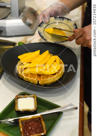 the cook putting fresh sliced mango fruit on baked belgian waffles with tongs the cook putting fresh sliced mango fruit on baked belgian waffles with tongs 107214938