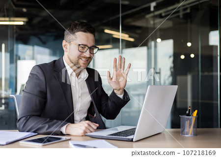 Smiling young businessman working in office, talking on video call on laptop, waving hand to camera. 107218169
