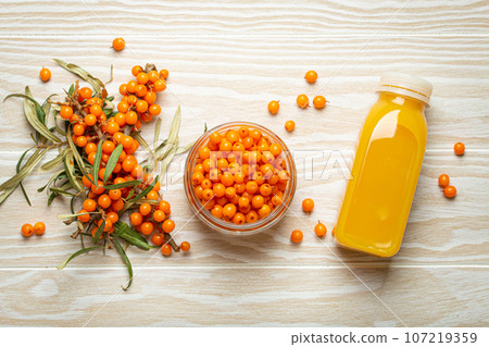 Sea buckthorn healthy drink in bottle, ripe berries in glass jar and branches with leaves top view on white wooden rustic background, great for skin, heart, vessels and immune system. 107219359