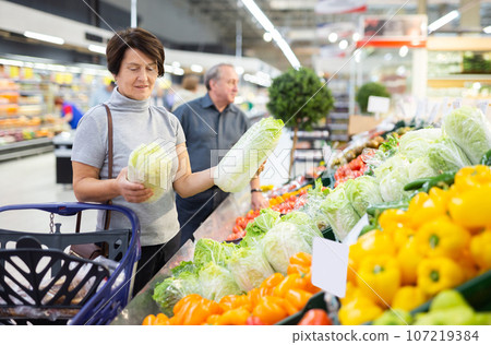 Elderly woman examines china cabbage in vegetable section to choose juiciest cabbage 107219384