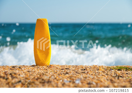 Cosmetic bottle on sandy beach with stormy wave of foam, blank label for mockup branding. Sunscreen cosmetics. Body lotion or cream with UV protection Cosmetic bottle on sandy beach with stormy wave of foam, blank label for mockup branding. Sunscreen cosmetics. Body lotion or cream with UV protection 107219815