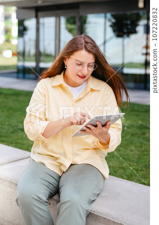 Beautiful smiling female student using tablet outdoors. Happy cheerful young woman siting on the bench on city street, Urban lifestyle concept. Traveler, Online education, business or shopping 107220832