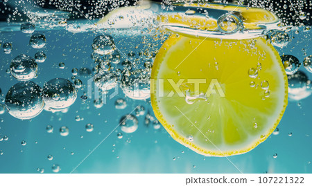 Underwater of freshly squeezed sweetened lemonade which raw lemons slice falling in soda water against dark blue or black background. Close up lemonade or Highball lemon cocktail cold refreshing drink Underwater of freshly squeezed sweetened lemonade which raw lemons slice falling in soda water against dark blue or black background. Close up lemonade or Highball lemon cocktail cold refreshing drink 107221322