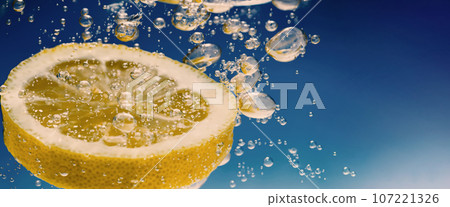 Underwater of freshly squeezed sweetened lemonade which raw lemons slice falling in soda water against dark blue or black background. Close up lemonade or Highball lemon cocktail cold refreshing drink Underwater of freshly squeezed sweetened lemonade which raw lemons slice falling in soda water against dark blue or black background. Close up lemonade or Highball lemon cocktail cold refreshing drink 107221326