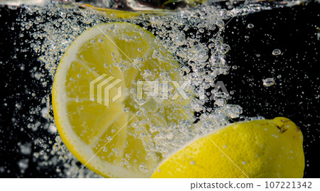 Underwater of freshly squeezed sweetened lemonade which raw lemons slice falling in soda water against dark blue or black background. Close up lemonade or Highball lemon cocktail cold refreshing drink Underwater of freshly squeezed sweetened lemonade which raw lemons slice falling in soda water against dark blue or black background. Close up lemonade or Highball lemon cocktail cold refreshing drink 107221342
