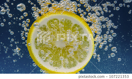 Underwater of freshly squeezed sweetened lemonade which raw lemons slice falling in soda water against dark blue or black background. Close up lemonade or Highball lemon cocktail cold refreshing drink Underwater of freshly squeezed sweetened lemonade which raw lemons slice falling in soda water against dark blue or black background. Close up lemonade or Highball lemon cocktail cold refreshing drink 107221353