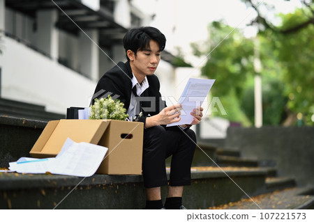 Stressed male office worker reading bad news notification about dismissal, sitting on stairs of building with cardboard box of belongings Stressed male office worker reading bad news notification about dismissal, sitting on stairs of building with cardboard box of belongings 107221573