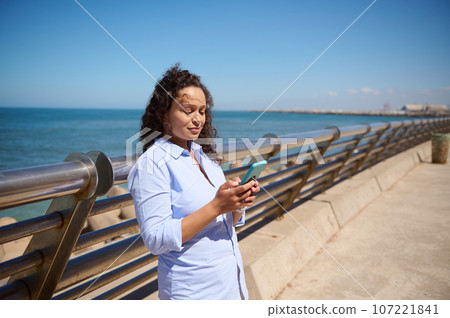 Beautiful ethnic woman in blue casual shirt checking mobile app on her smartphone, smiling, enjoying beautiful sunny day 107221841