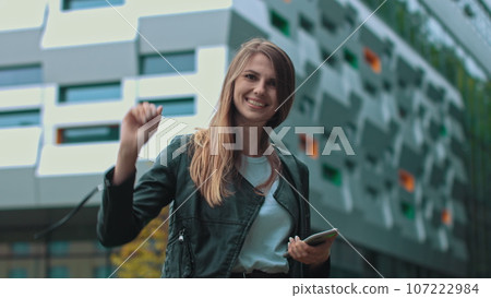 Good looking girl with Long Brown Hair Looking at Her phone Typing and Smiling. Enormous industrial Building at the Background. Green Bushes and Trees. Smart clothes. Natural makeup Good looking girl with Long Brown Hair Looking at Her phone Typing and Smiling. Enormous industrial Building at the Background. Green Bushes and Trees. Smart clothes. Natural makeup 107222984