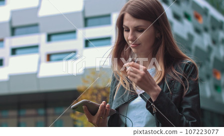 Attractive girl, student, in modern clothes Uses a mobile phone to listen to good mood music, music lover puts a favorite song, happy woman uses roaming, high speed connection 5G. Attractive girl, student, in modern clothes Uses a mobile phone to listen to good mood music, music lover puts a favorite song, happy woman uses roaming, high speed connection 5G. 107223077
