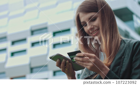 Smiling young female customer holding credit card and smartphone, on modern home background. Happy women shoppers using instant simple mobile payments while shopping online. Electronic banking service Smiling young female customer holding credit card and smartphone, on modern home background. Happy women shoppers using instant simple mobile payments while shopping online. Electronic banking service 107223092