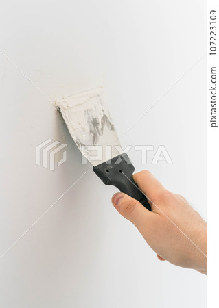 Male using spatula and plastering of wall with white fresh finishing putty. Closeup. Repair work of home. Side view. 107223109