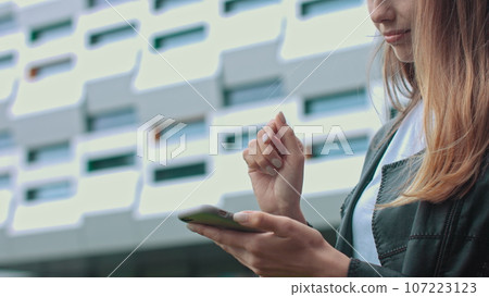 Young attractive girl in stylish clothes on the background of a modern building, looks at the phone and enters a message. Touch in the air with your index finger. Online shopping. The girl makes a 107223123
