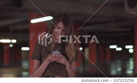 Relaxed young woman using smartphone surfing social media, checking news, playing mobile games or texting messages standing in the supermarket parking lot 107223256