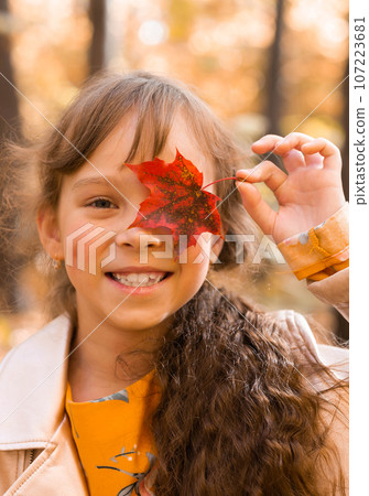 Close portrait of a little girl. She covers her eye with a maple autumn leaf. Fall season and children concept. 107223681
