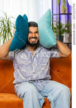Neighbors repair work, irritated man by loud music at home cover ears with pillows annoyed by noise Neighbors repair work, irritated man by loud music at home cover ears with pillows annoyed by noise 107223754