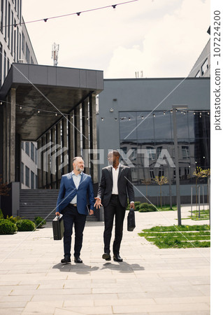 Confident businesspeople standing in front of modern office building. Black and caucasian men talking. Banking and financial market concept. Confident businesspeople standing in front of modern office building. Black and caucasian men talking. Banking and financial market concept. 107224200